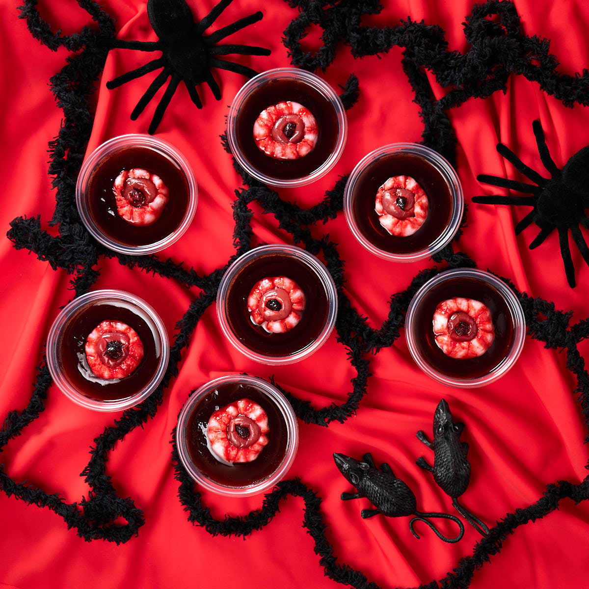 Rum and coke jello shots with eyeballs spread on a red table cloth with plastic spiders and rats.