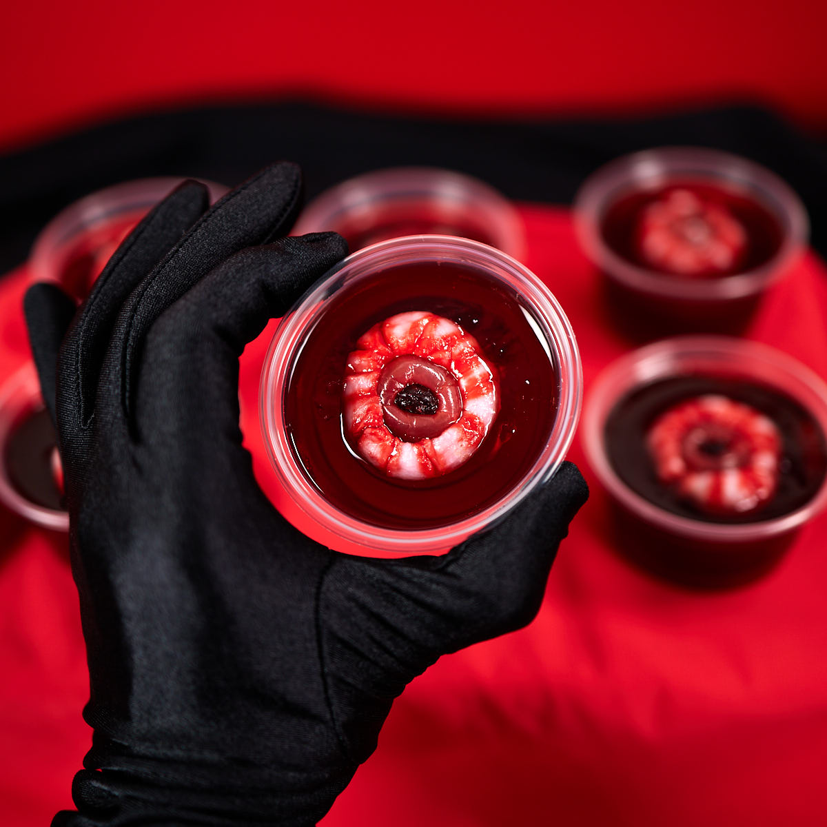 Gory red jello shot with bloody eyeball held in a black gloved hand.