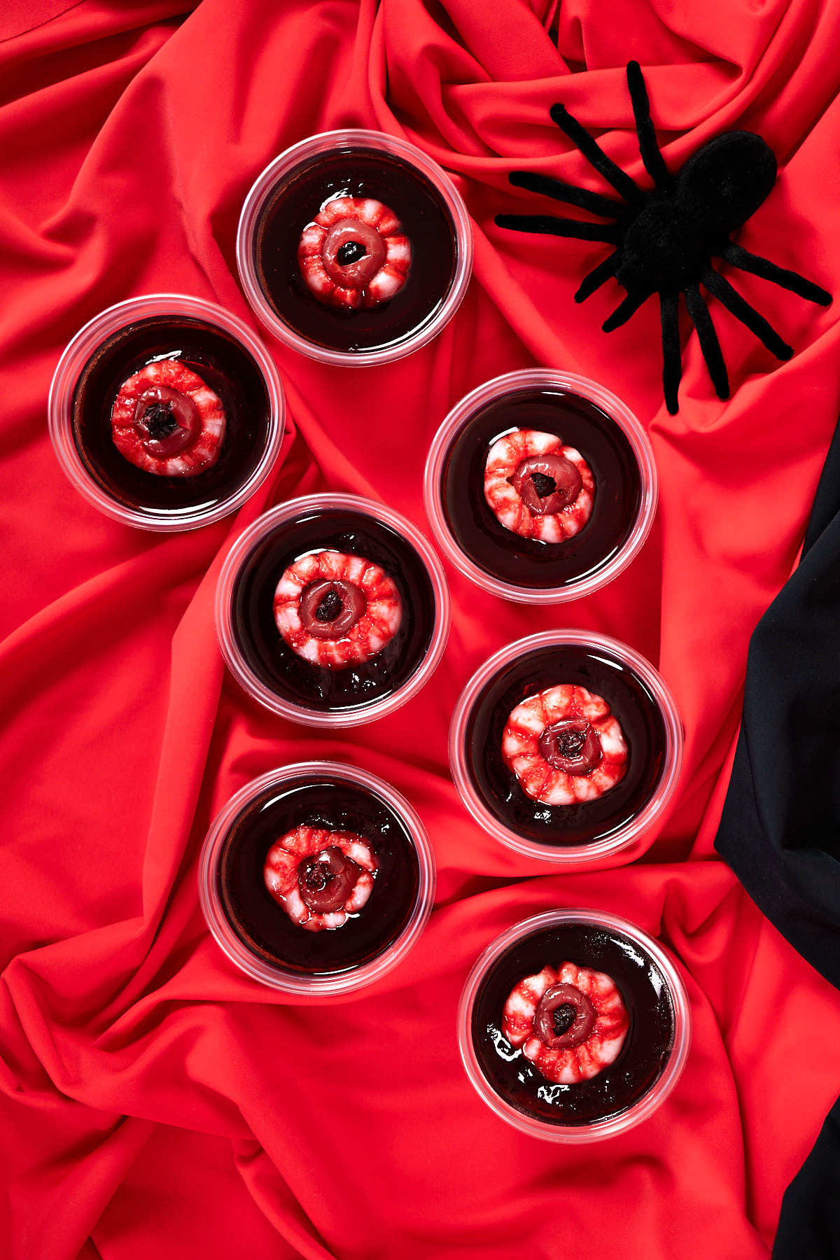 Halloween rum and coke jello shots with eyeballs spread over a red cloth on a table.