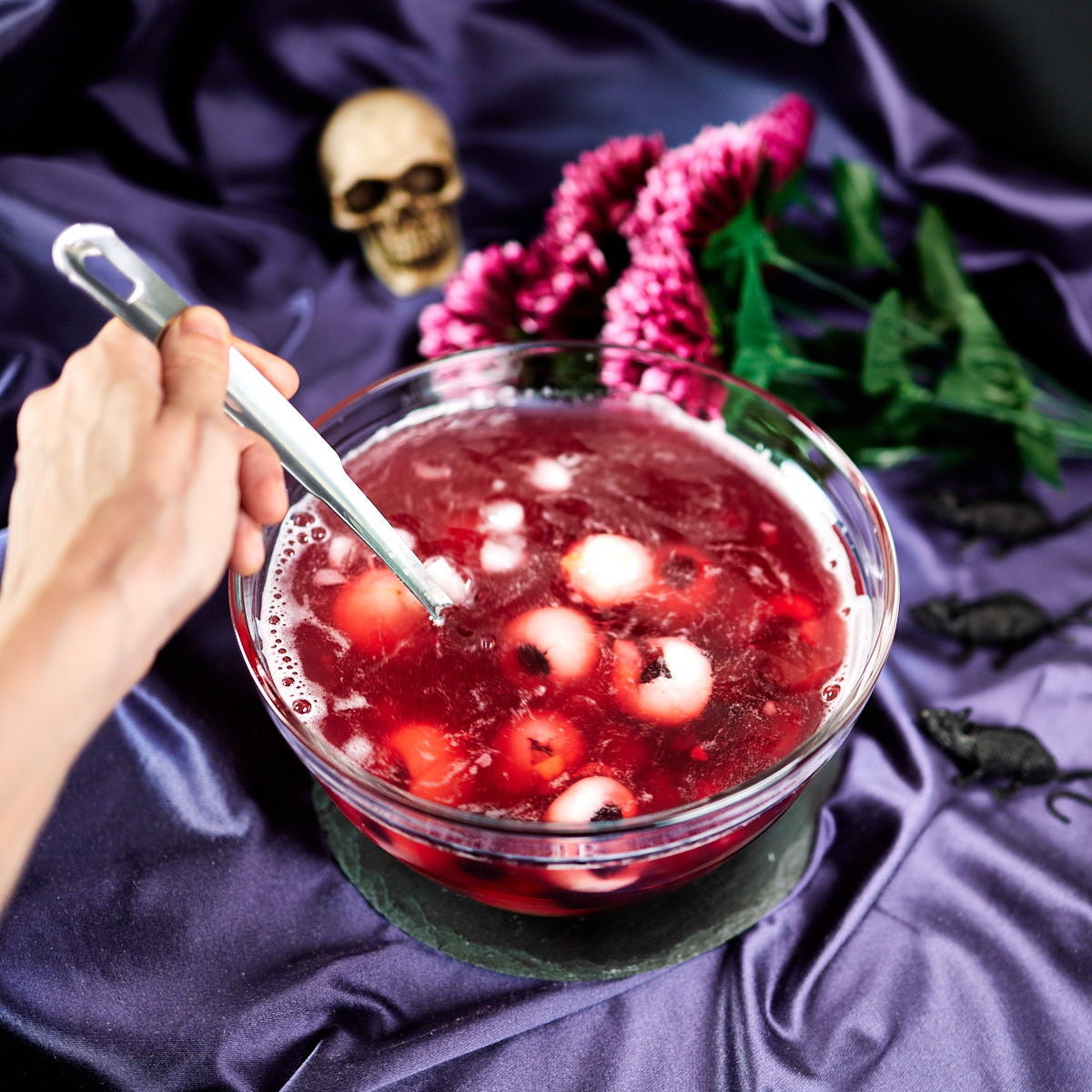 Mixing a glass bowl of purplish colored punch with lychee eyeballs, Halloween decoration in the background.